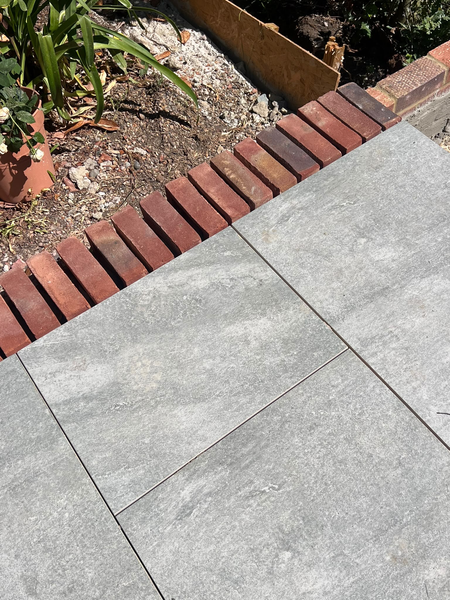 A hand-laid brick soldier course edging grey porcelain paving, with a planting bed and terracotta pot alongside.